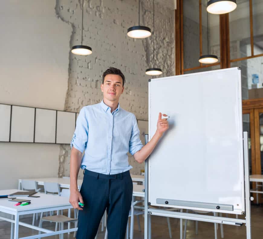 A man in a light blue shirt stands next to a blank whiteboard in a modern classroom, pointing at the board with a marker. Tables, chairs, and pendant lights are visible in the background.